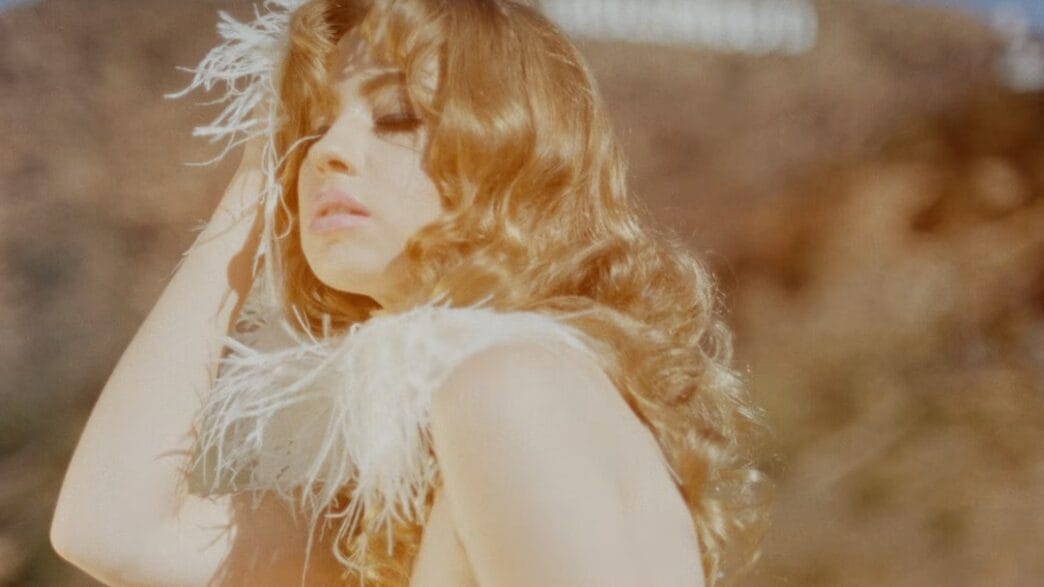 Sunlit portrait of a blonde woman with wavy hair, wearing a pale pink strapless dress and a feathered accessory, posing outdoors.