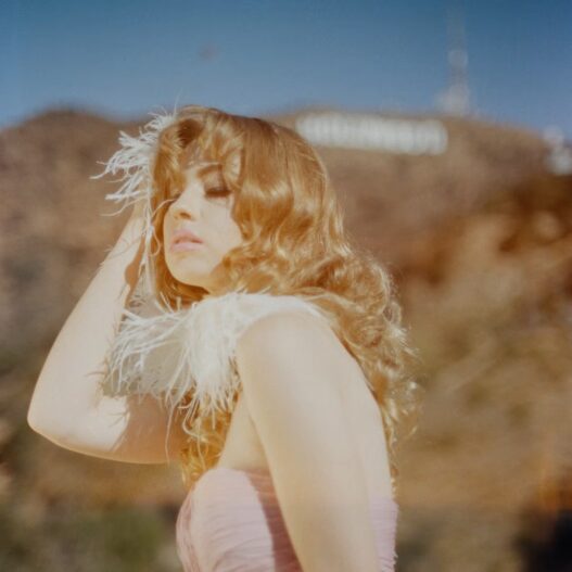 Sunlit portrait of a blonde woman with wavy hair, wearing a pale pink strapless dress and a feathered accessory, posing outdoors.
