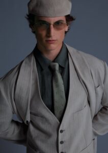 Portrait of a young man in a light gray suit, vest, and paisley tie, wearing a cap and tinted glasses.