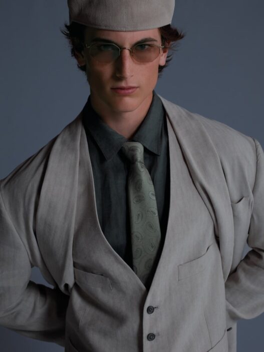 Portrait of a young man in a light gray suit, vest, and paisley tie, wearing a cap and tinted glasses.