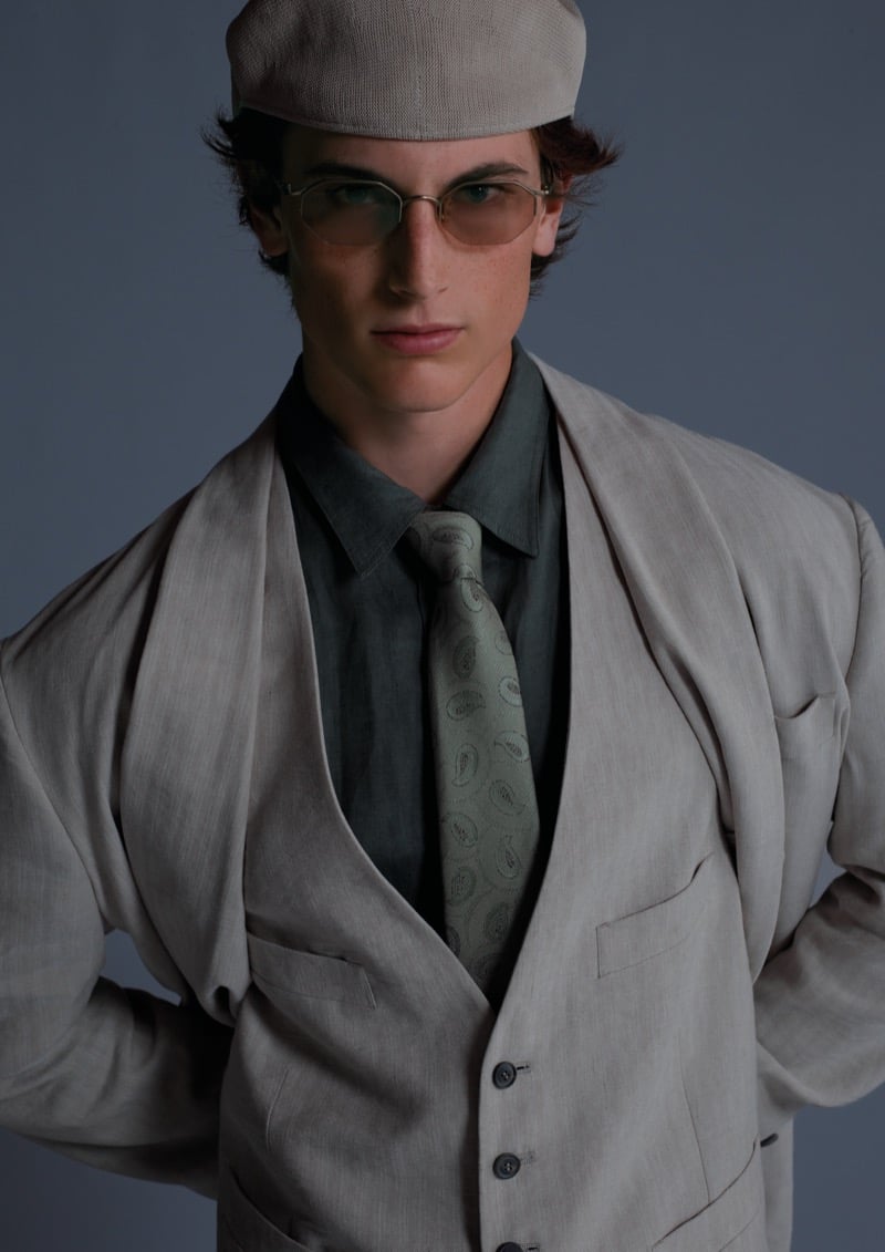 Portrait of a young man in a light gray suit, vest, and paisley tie, wearing a cap and tinted glasses.