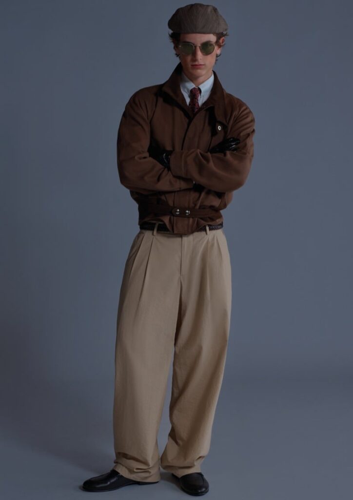 Man in a vintage 1940s detective outfit: brown trench coat, tan pleated trousers, cap, sunglasses, arms crossed.