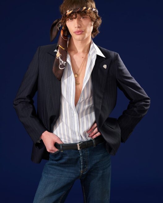 Male fashion model wearing a dark pinstripe blazer over an unbuttoned striped shirt, jeans, and a brown scarf tied around his head against a dark blue backdrop.