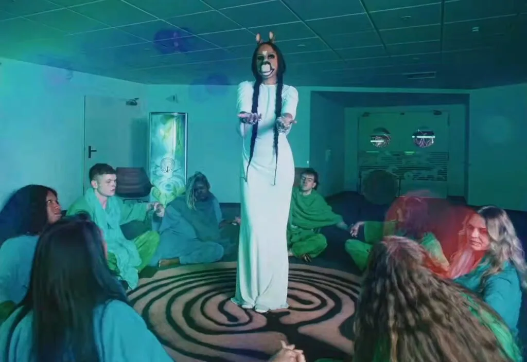 Center-stage performer in a white gown with braided hair and a donkey mask, standing on a spiral rug as a seated audience watches in a dim blue-green room.