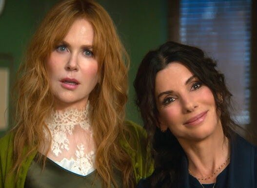 Two women posing indoors side by side, the red-haired one in a lace high-neck top and green cardigan, the other in a dark blazer, both smiling.