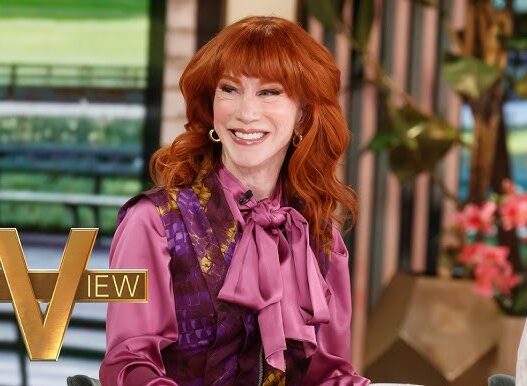 Smiling red-haired woman in a pink blouse and purple vest sits at a talk-show set with the 'THE VIEW' logo visible in the foreground.