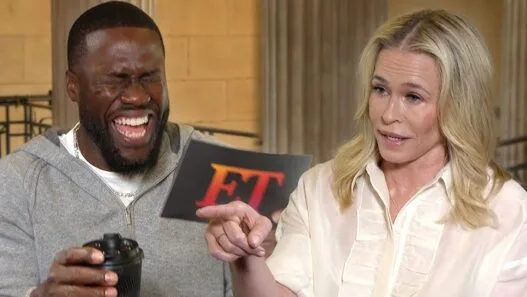Man laughing with a camera in hand, while a woman points to a card with red 'ET' letters during an interview.