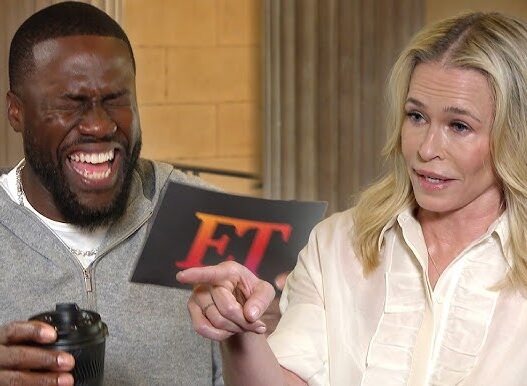 Man laughing with a camera in hand, while a woman points to a card with red 'ET' letters during an interview.