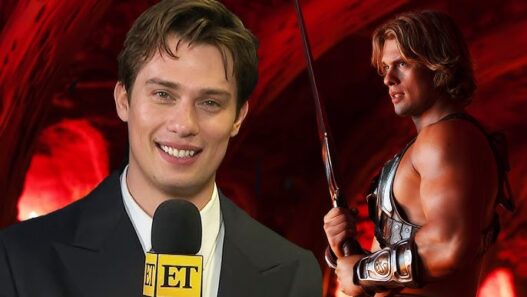 Entertainment Tonight reporter speaking into a mic in foreground, with a muscular warrior in armor and sword in a red-lit background set.