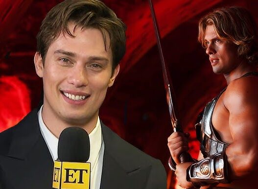 Entertainment Tonight reporter speaking into a mic in foreground, with a muscular warrior in armor and sword in a red-lit background set.