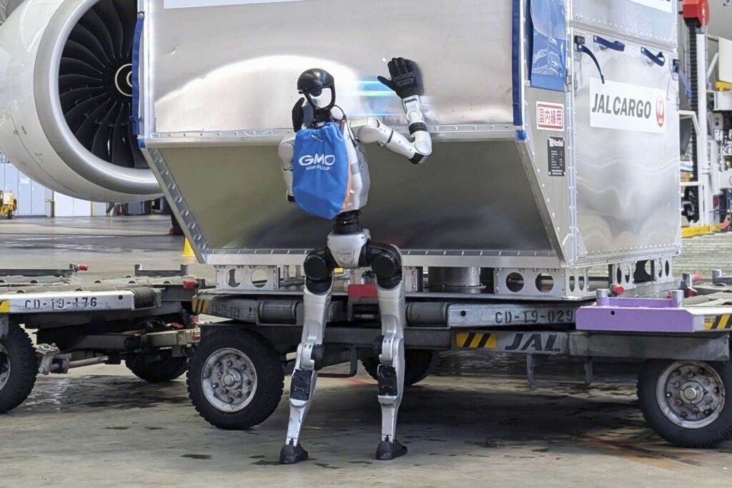 Humanoid robot in a blue vest stands under a large silver JAL Cargo container on a loading pallet in a hangar.