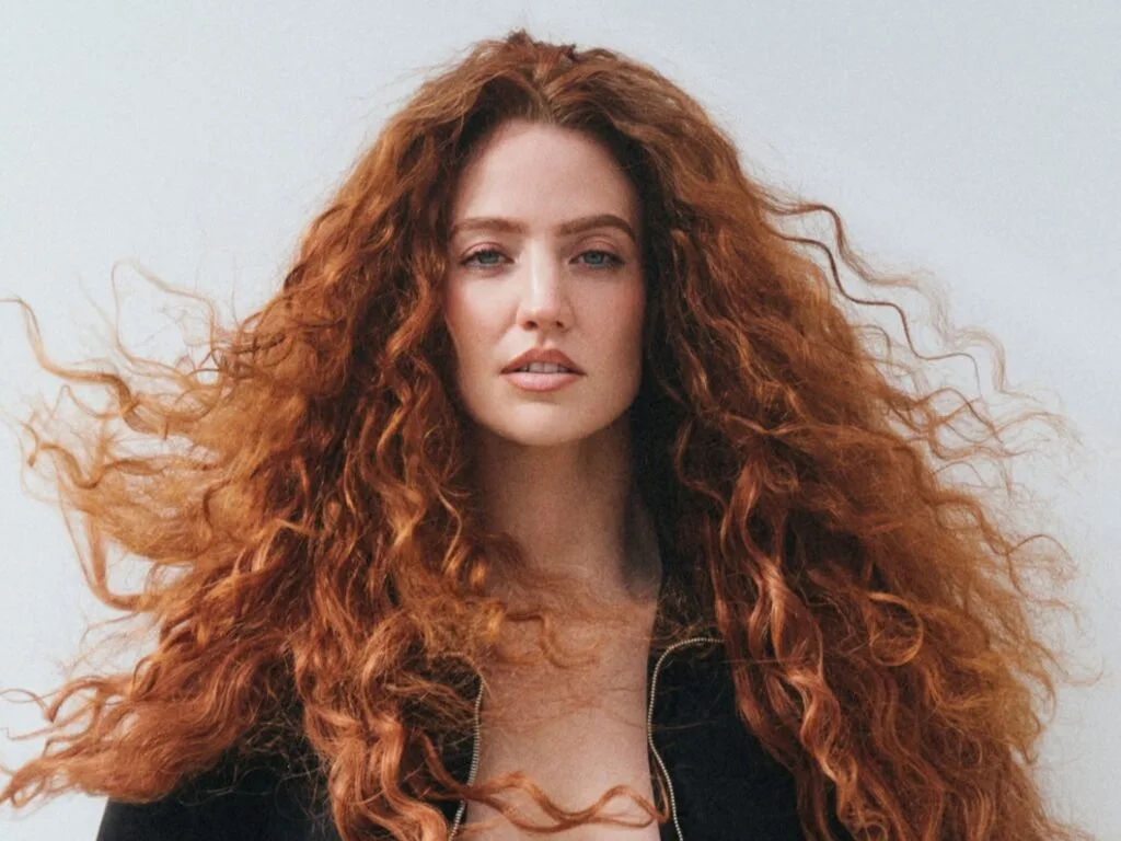 Woman with long, voluminous red curls facing the camera, wearing a dark jacket against a pale background.