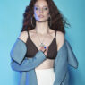 Portrait of a woman with curly red hair against a blue backdrop, wearing a brown bralette, a light blue coat, large hoop earrings, and a silver leaf necklace.