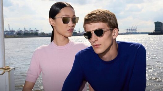 Two people wearing sunglasses by a sunny waterfront, posing for a photo; man in a blue shirt, woman in a pink top with slicked-back hair.