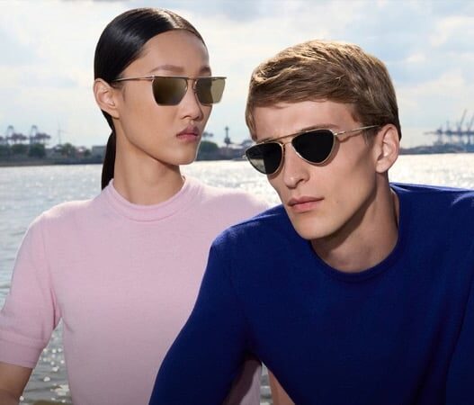 Two people wearing sunglasses by a sunny waterfront, posing for a photo; man in a blue shirt, woman in a pink top with slicked-back hair.