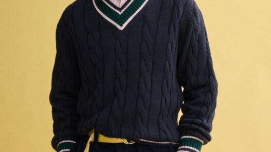 Model Klevi Rybak wearing the J.Press x Alex Mill navy cable‑knit cricket sweater. Man wearing a navy cable-knit V-neck sweater with green and white trim over a pink shirt, paired with navy shorts and a yellow belt against a yellow backdrop