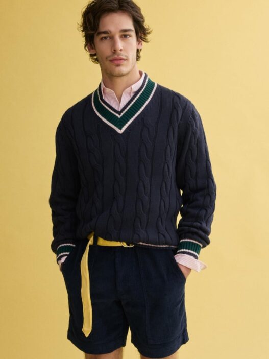 Model Klevi Rybak wearing the J.Press x Alex Mill navy cable‑knit cricket sweater. Man wearing a navy cable-knit V-neck sweater with green and white trim over a pink shirt, paired with navy shorts and a yellow belt against a yellow backdrop