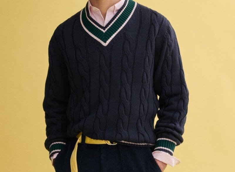 Model Klevi Rybak wearing the J.Press x Alex Mill navy cable‑knit cricket sweater. Man wearing a navy cable-knit V-neck sweater with green and white trim over a pink shirt, paired with navy shorts and a yellow belt against a yellow backdrop