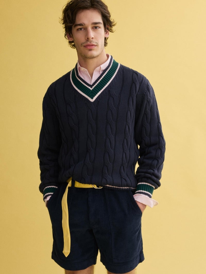 Model Klevi Rybak wearing the J.Press x Alex Mill navy cable‑knit cricket sweater. Man wearing a navy cable-knit V-neck sweater with green and white trim over a pink shirt, paired with navy shorts and a yellow belt against a yellow backdrop