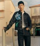 Young man wearing a dark denim jacket with a large blue floral print over a white shirt, posing outdoors near buildings.