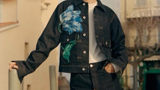 Young man wearing a dark denim jacket with a large blue floral print over a white shirt, posing outdoors near buildings.