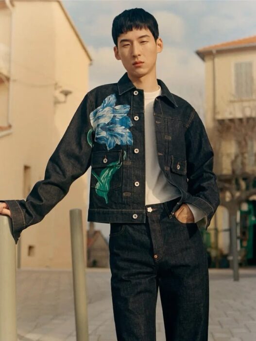 Young man wearing a dark denim jacket with a large blue floral print over a white shirt, posing outdoors near buildings.
