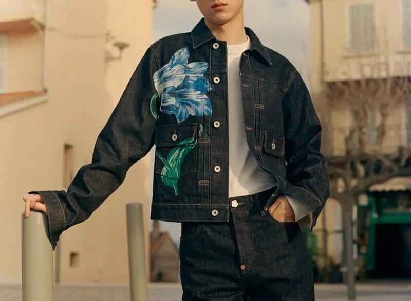 Young man wearing a dark denim jacket with a large blue floral print over a white shirt, posing outdoors near buildings.