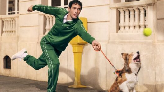 Man in a green tracksuit releases a throw as a dog on a leash jumps to catch a tennis ball on an urban sidewalk near a beige building with a yellow post nearby. Lacoste unveils its revived “Life Is a Beautiful Sport” campaign directed by Fredrik Bond.