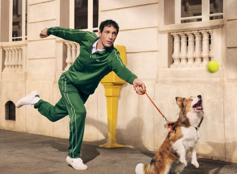 Man in a green tracksuit releases a throw as a dog on a leash jumps to catch a tennis ball on an urban sidewalk near a beige building with a yellow post nearby. Lacoste unveils its revived “Life Is a Beautiful Sport” campaign directed by Fredrik Bond.