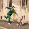 Man in a green tracksuit releases a throw as a dog on a leash jumps to catch a tennis ball on an urban sidewalk near a beige building with a yellow post nearby. Lacoste unveils its revived “Life Is a Beautiful Sport” campaign directed by Fredrik Bond.