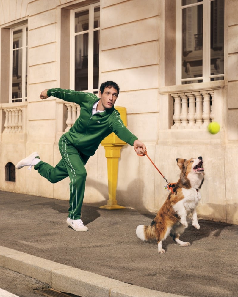 Man in a green tracksuit releases a throw as a dog on a leash jumps to catch a tennis ball on an urban sidewalk near a beige building with a yellow post nearby. Lacoste unveils its revived “Life Is a Beautiful Sport” campaign directed by Fredrik Bond.