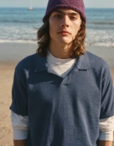 Young person with shoulder-length hair wearing a purple knit beanie and a blue polo over a white shirt, standing on a sandy beach.