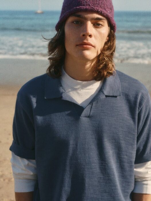 Young person with shoulder-length hair wearing a purple knit beanie and a blue polo over a white shirt, standing on a sandy beach.
