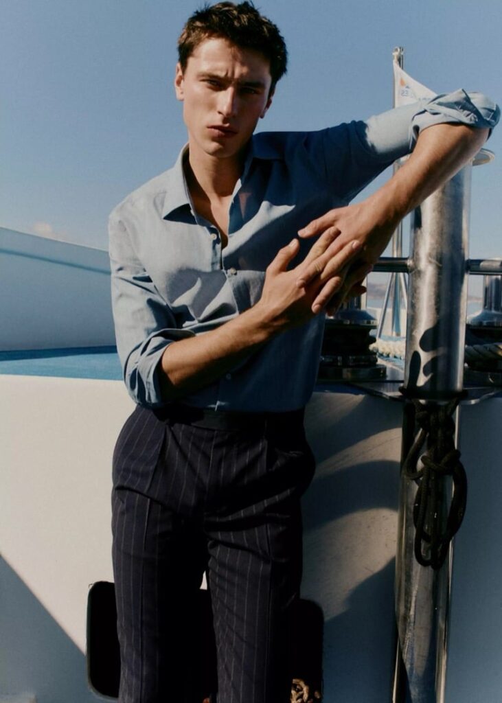 Young man leaning on a boat railing in a light blue shirt and dark pinstripe trousers, against a clear blue sky.