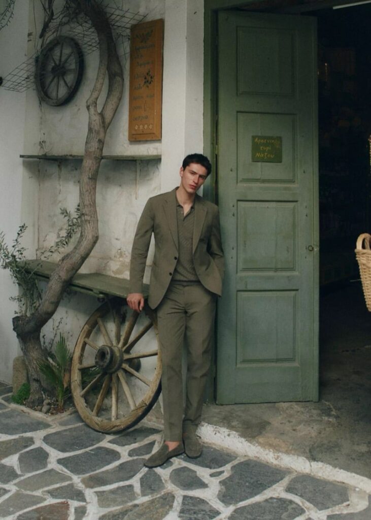 Man in an olive suit leaning against a green door of a rustic storefront with a wooden wagon wheel and stone pavement behind him to the left index finger in pocket.