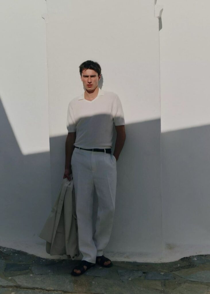 Young man in white polo and light pants stands against a white wall, holding a light jacket and wearing sandals, with shadow across his torso