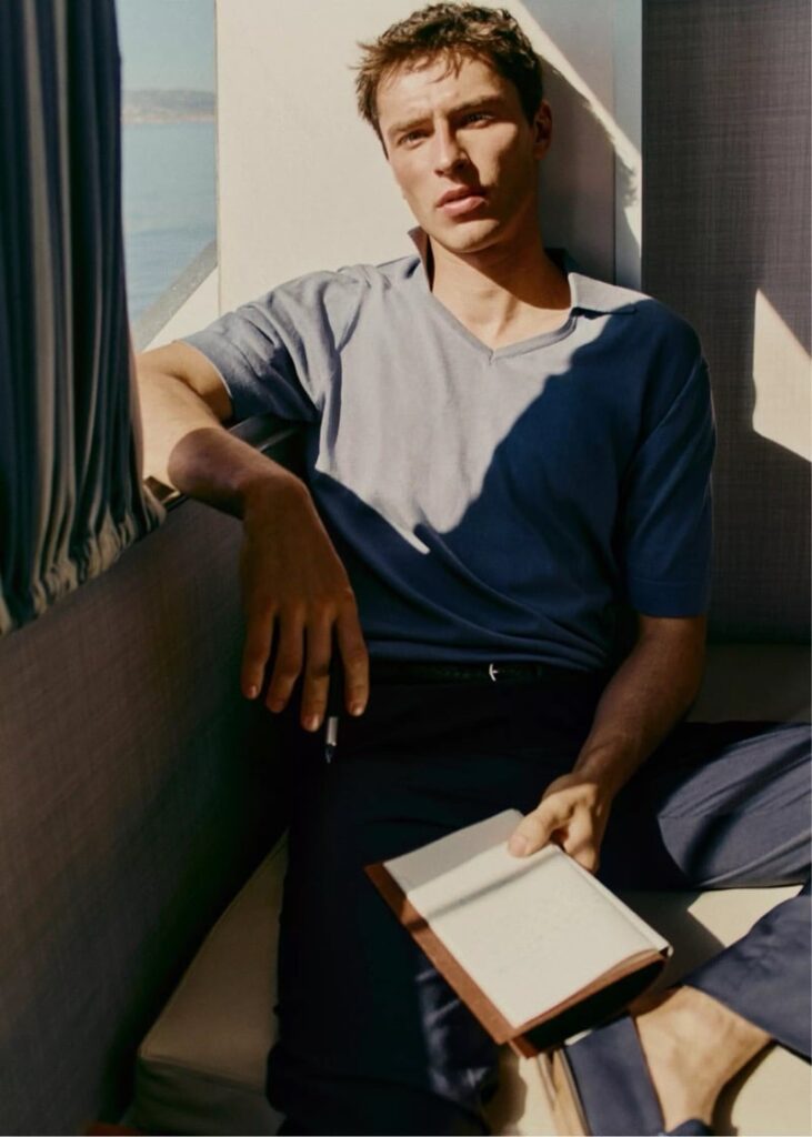 Young man sits on a couch by a window, holding a closed notebook and looking at the camera in sunlight.