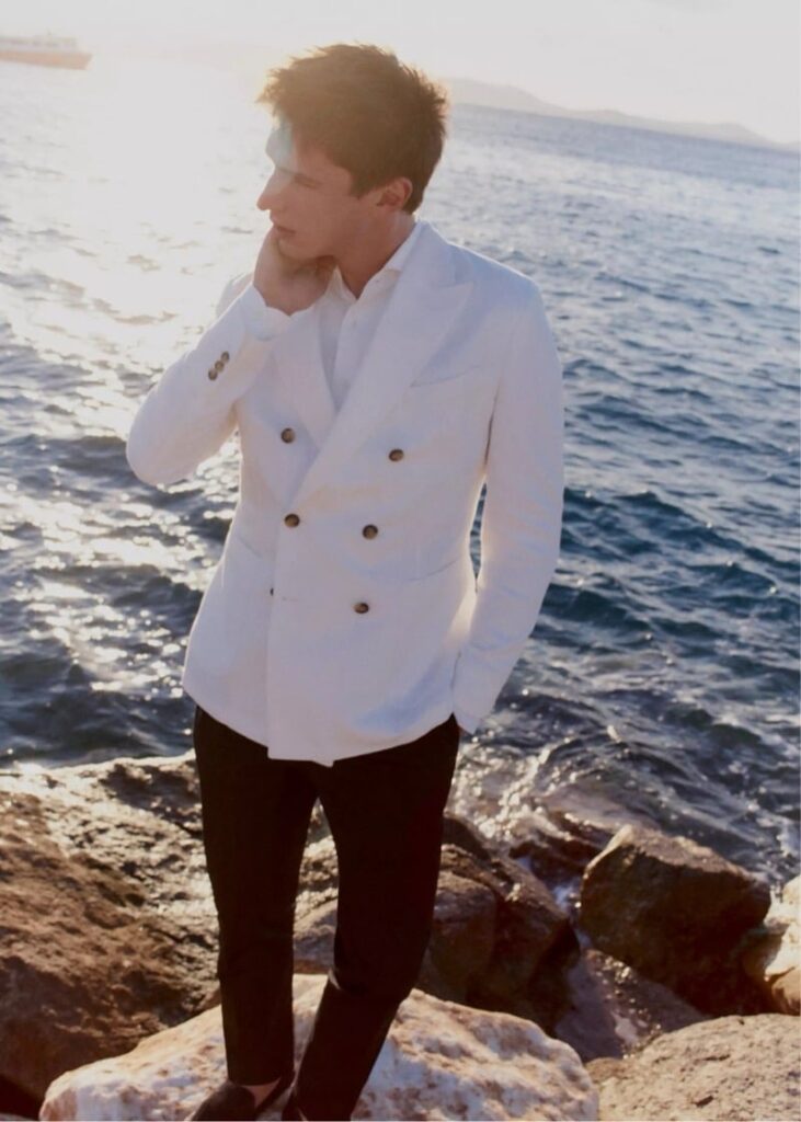 Man standing on rocks by the sea at sunset, wearing a white double‑breasted blazer and dark pants, looking to the side with hand near his chin.