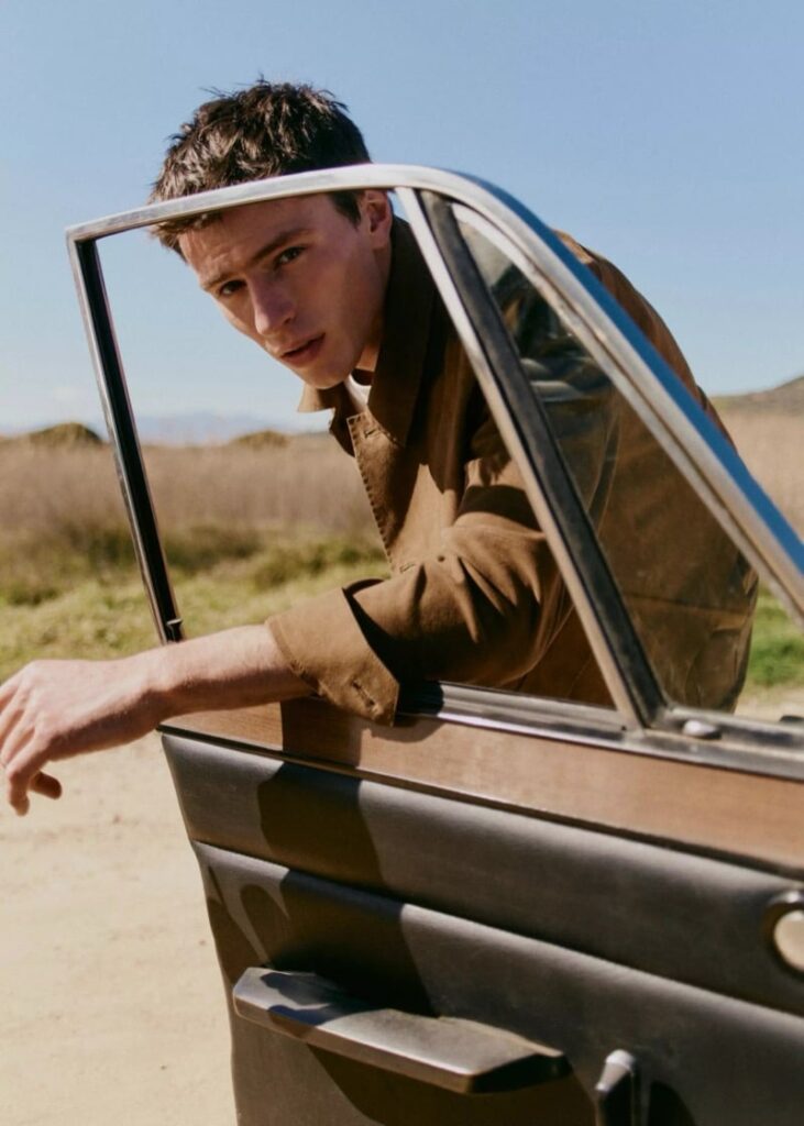 Young man leans out of a vintage car window, looking toward the camera in a desert landscape.