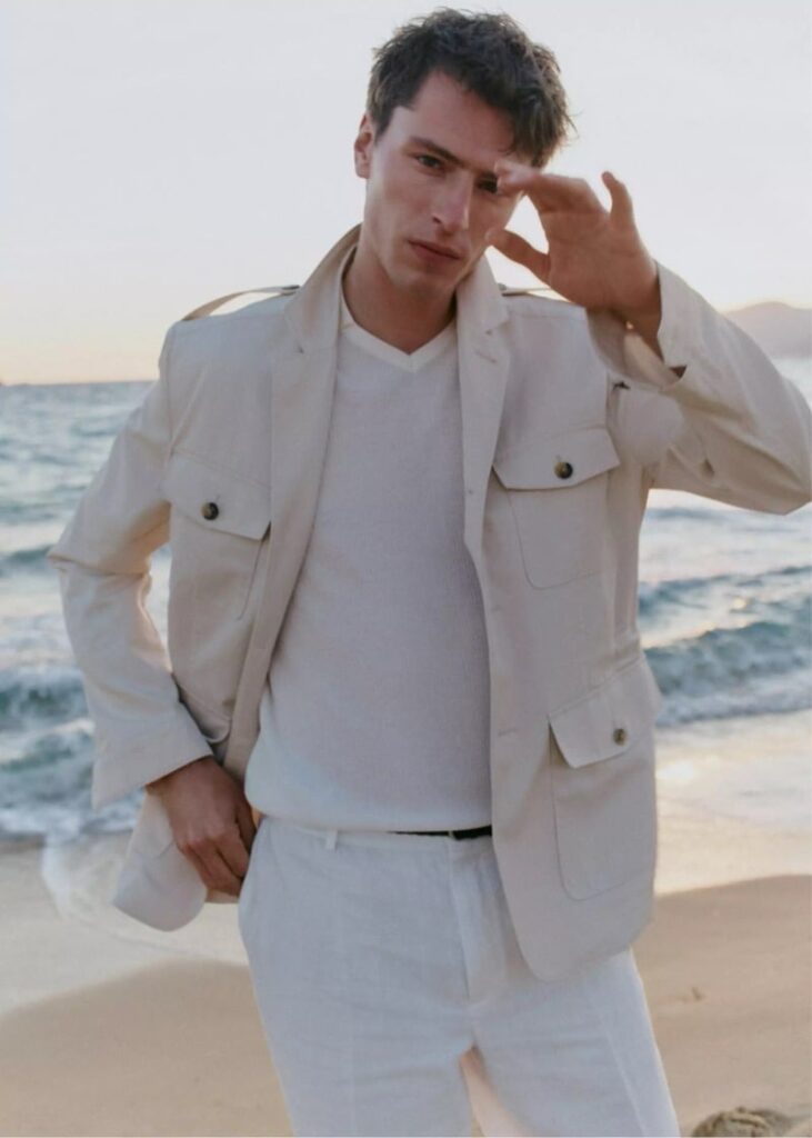 Man posing on a beach in a light beige jacket and white outfit, shielding his eyes from the sun with one hand.