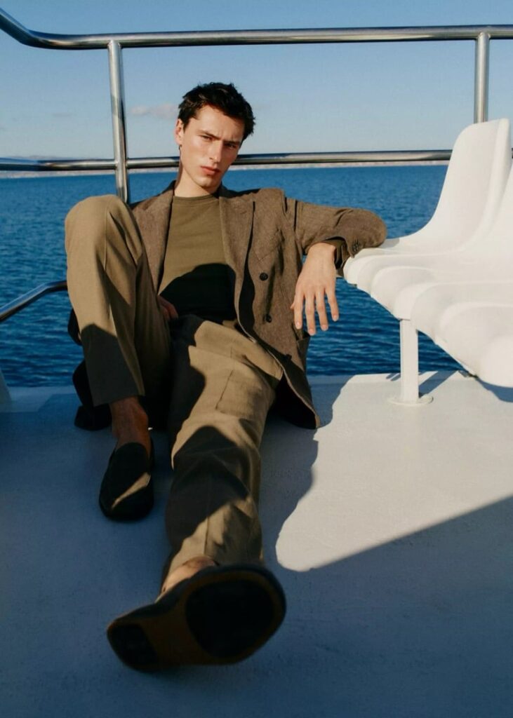 Young man in a brown suit reclining on a boat deck with the ocean in the background