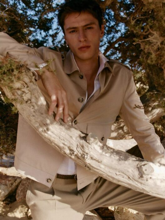 Young man in a beige suit and white shirt leans on a large branch outdoors among trees, looking at the camera.