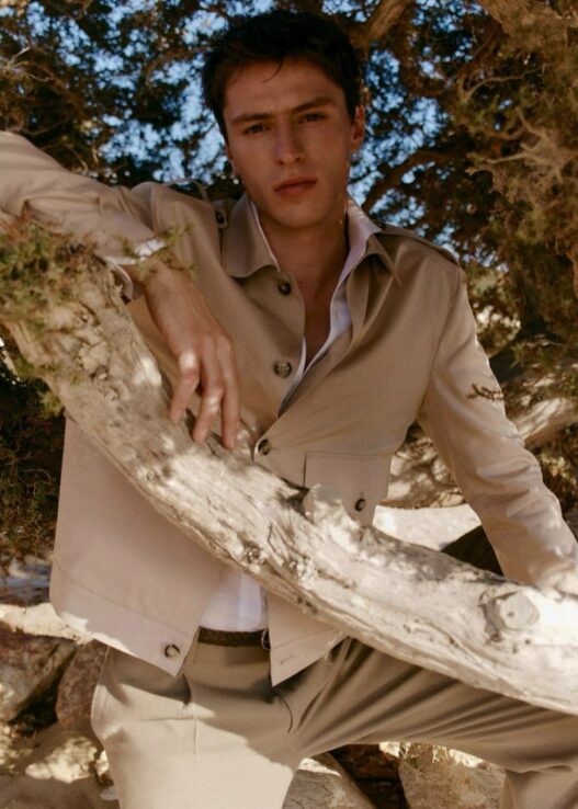 Young man in a beige suit and white shirt leans on a large branch outdoors among trees, looking at the camera.