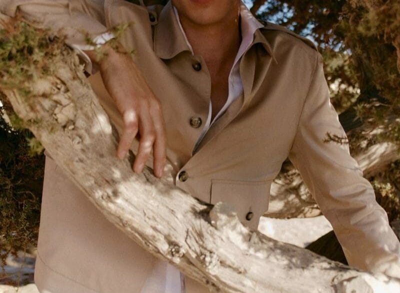 Young man in a beige suit and white shirt leans on a large branch outdoors among trees, looking at the camera.