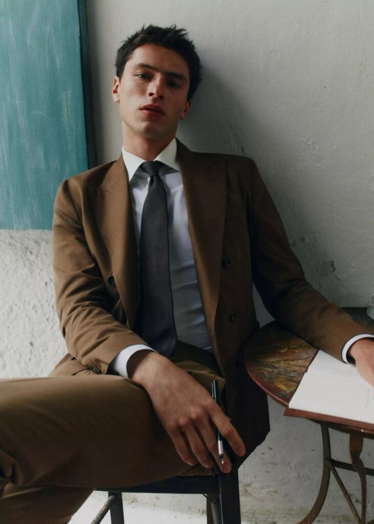 Young man in a brown suit sits leaning against a wall, holding a pen near a notebook on a small table.