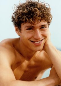 Young man with curly brown hair smiling, resting his head on his hand.
