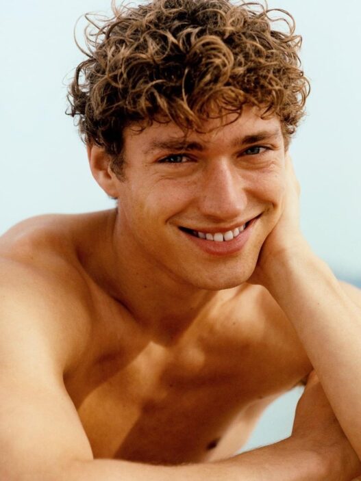 Young man with curly brown hair smiling, resting his head on his hand.