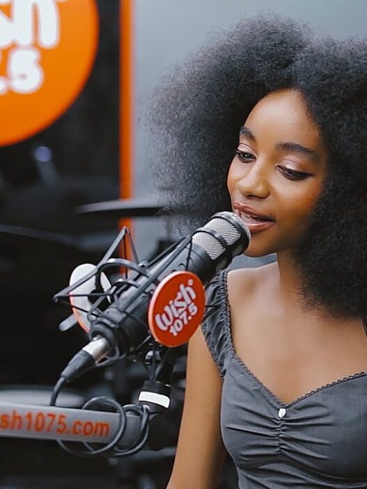 Woman with large curly hair speaks into a studio microphone at Wish 107.5, with the Wish logo visible.