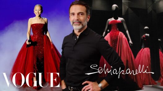 Man in a black polo posing in front of red ballgown mannequins on a Vogue/Schiaparelli backdrop.