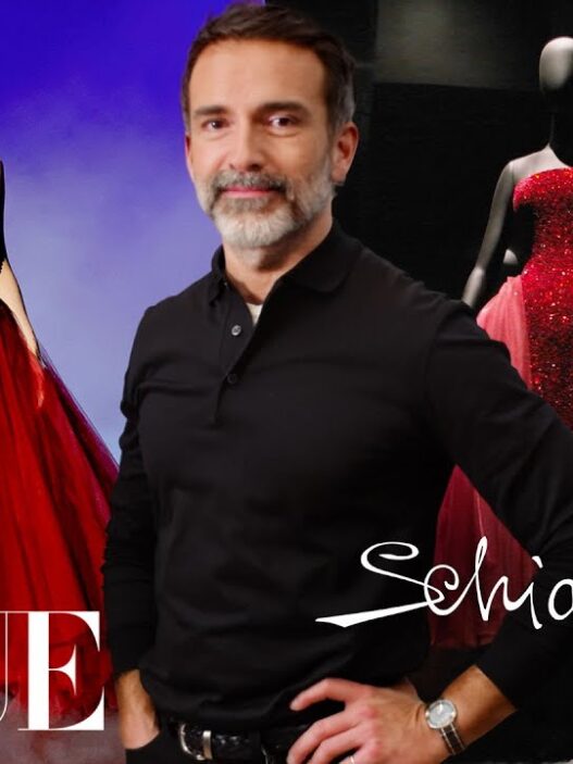 Man in a black polo posing in front of red ballgown mannequins on a Vogue/Schiaparelli backdrop.